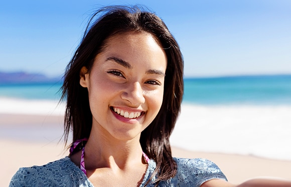 Se muestra una selfie de una mujer sonriendo, sujetando el dispositivo con la mano izquierda. Detrás de ella hay una vista brillante y borrosa de la playa y el cielo azul.