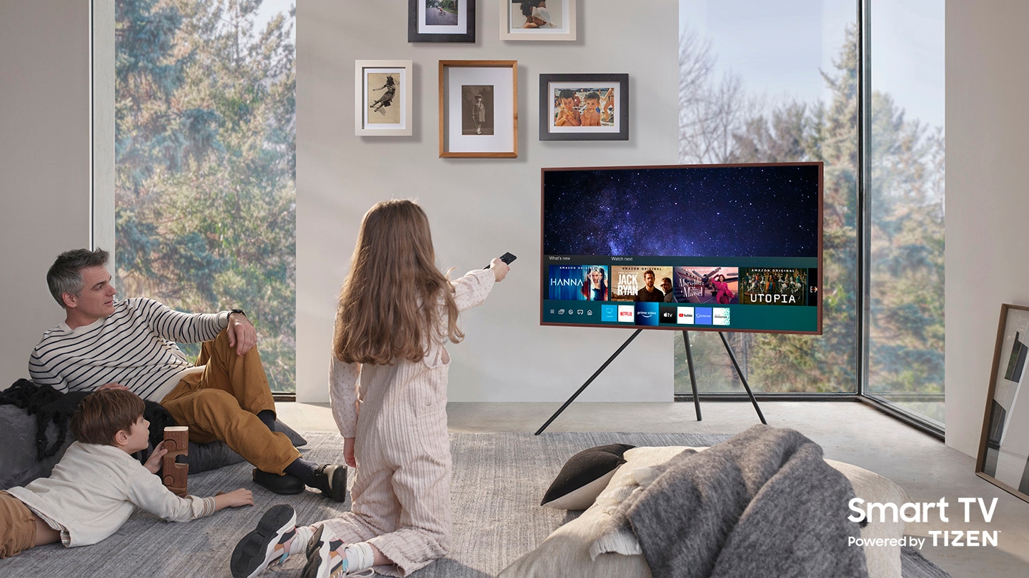 A father and his two children are enjoying The Frame which is mounted to Studio Stand. On the screen is the Universal Guide user interface menu which the girl is controlling with One Remote. The Smart TV Powered by Tizen logo is visible.