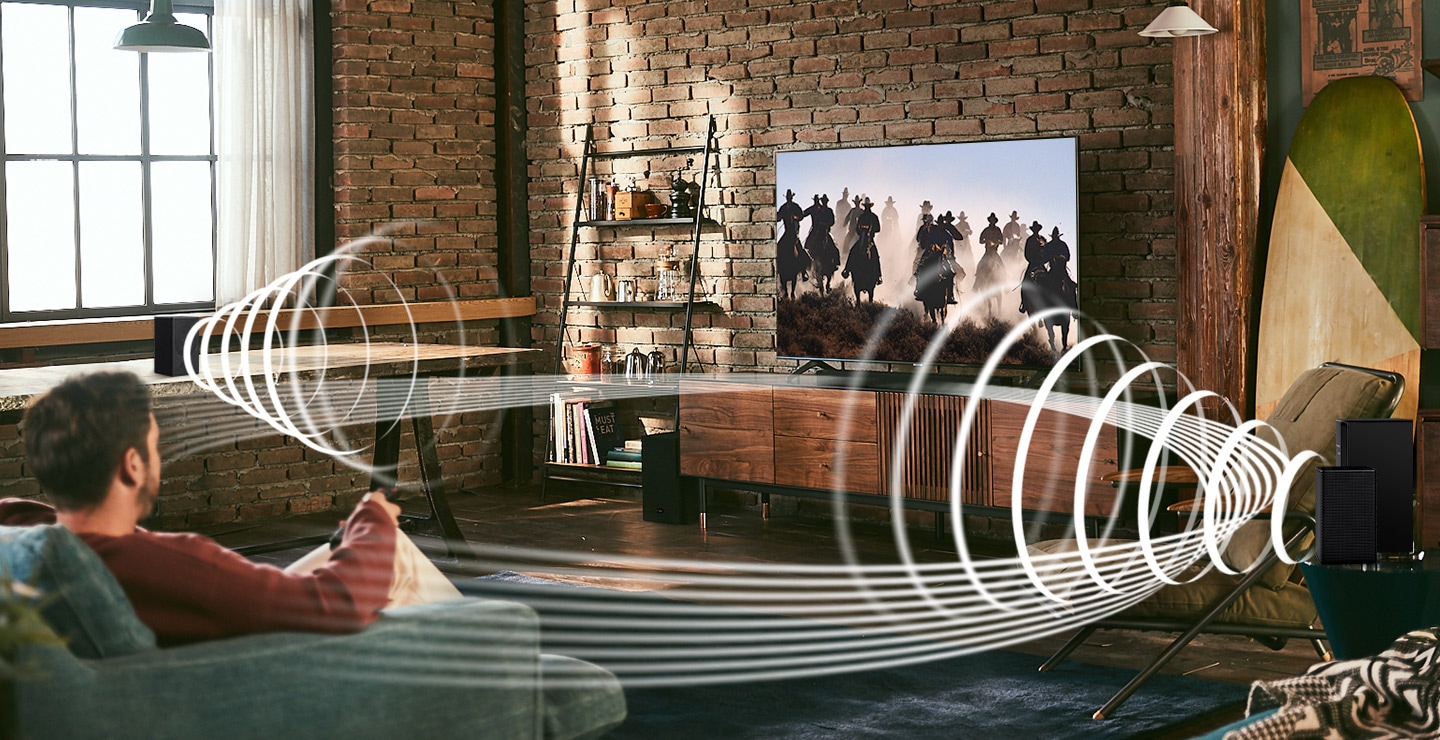 A man enjoys racing content on his TV. Soundwave graphics are playing from Samsung Wireless Rear Speaker Kit and Soundbar, demonstrating Wireless Surround Sound Compatible feature of Samsung soundbar.