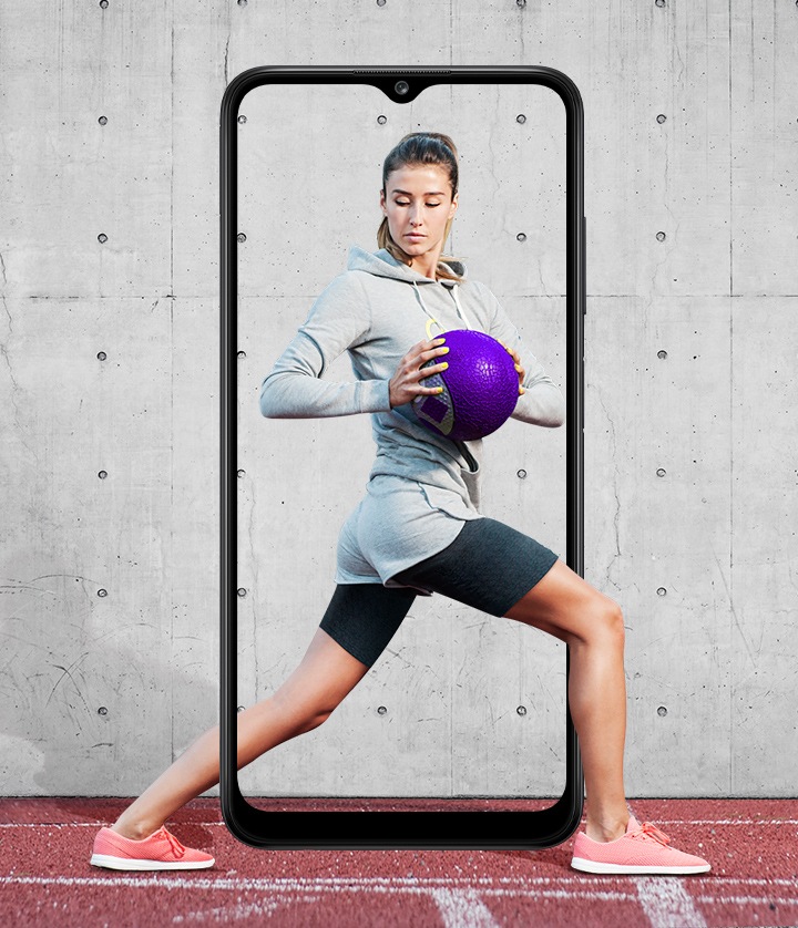 A woman is shown performing a standing torso twist with her legs in a lunge position while holding a purple medicine ball. The Galaxy A03s's slim bezel frames her entire body, with her feet and part of her calf outside it.