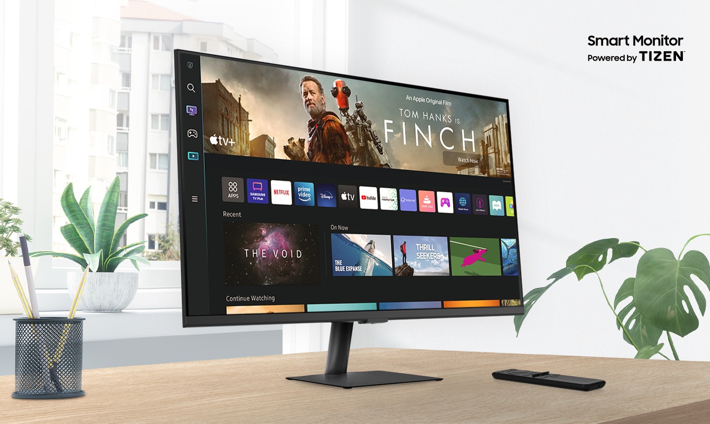 Smart Monitor M5 sits on top of a wooden desk in front of a window surrounded by plants. Above the monitor, texts read Smart Monitor Powered by Tizen. The screen shows an Apple TV app with a variety of content. A remote rests next to the monitor.