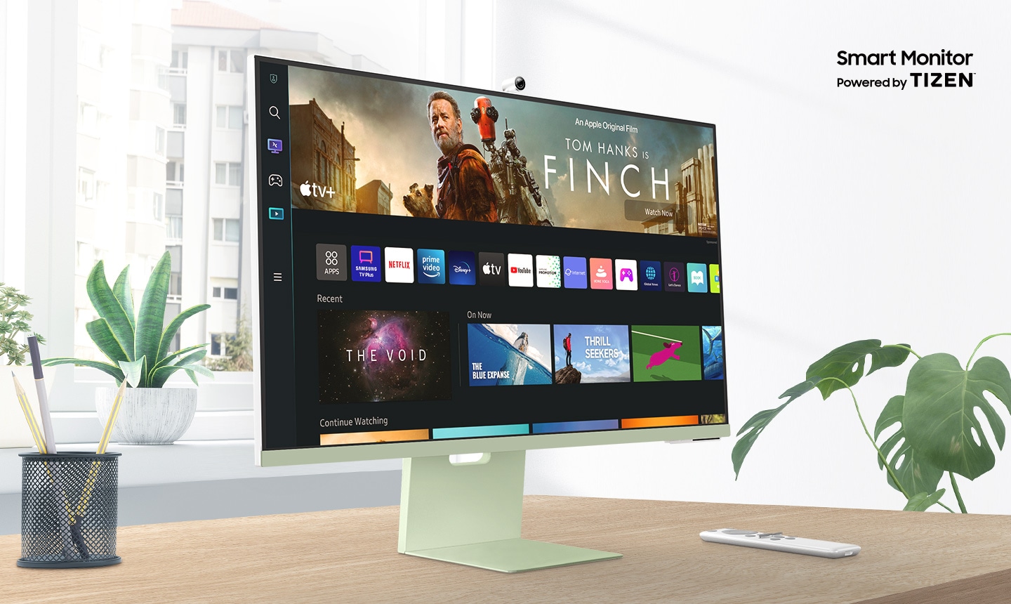 Samsung M8 32 inch smart monitor sits on top of a wooden desk in front of a window surrounded by plants. Above the monitor, texts read Smart Monitor Powered by Tizen. The screen shows an Apple TV app with a variety of content. A remote rests next to the monitor.