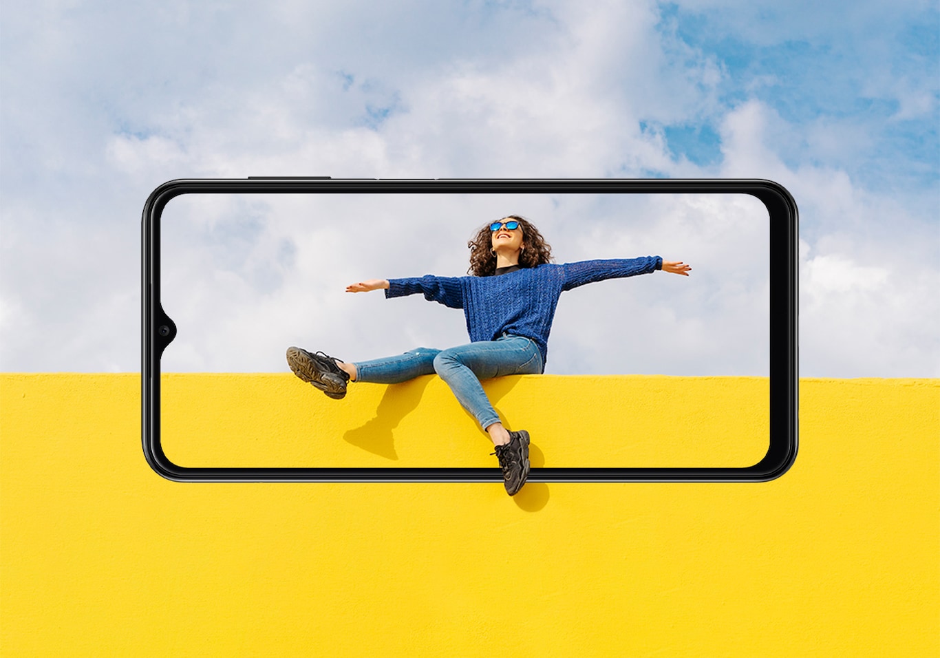 Keep a tab on the Samsung Galaxy A13 release date. Galaxy A13 seen from the front. A young woman sitting on yellow wall against cloudy sky looking up. Her legs and the wall and sky extend outside of the display.