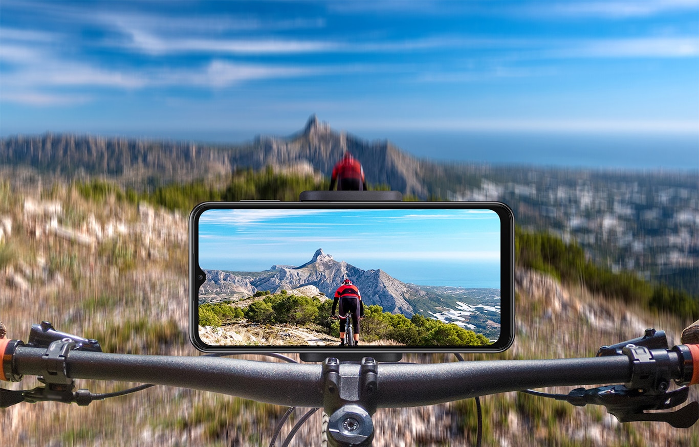 Shoot a stable video on Samsung Galaxy A23. A Galaxy A23 is mounted in landscape mode on a bicycle handlebar. In front, a blurry, mountainous terrain is shown and on-screen, a clear picture of the terrain as well as the rider in front is captured.