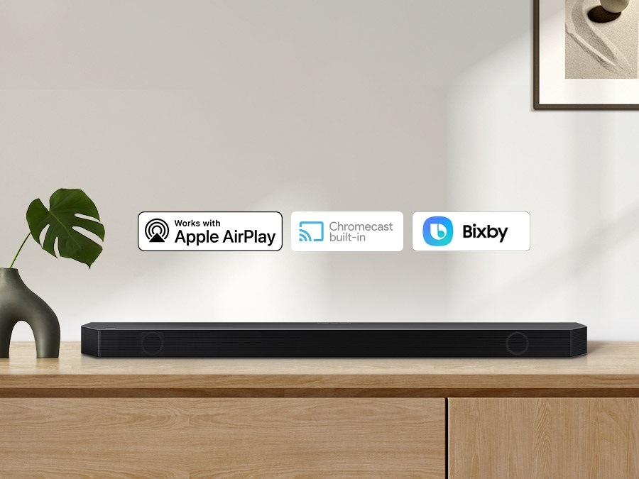 Apple AirPlay logo, Chromecast Built-in logo, and Ok Google logo can be seen along with Samsung Q930B soundbar which is sitting on living room cabinet.