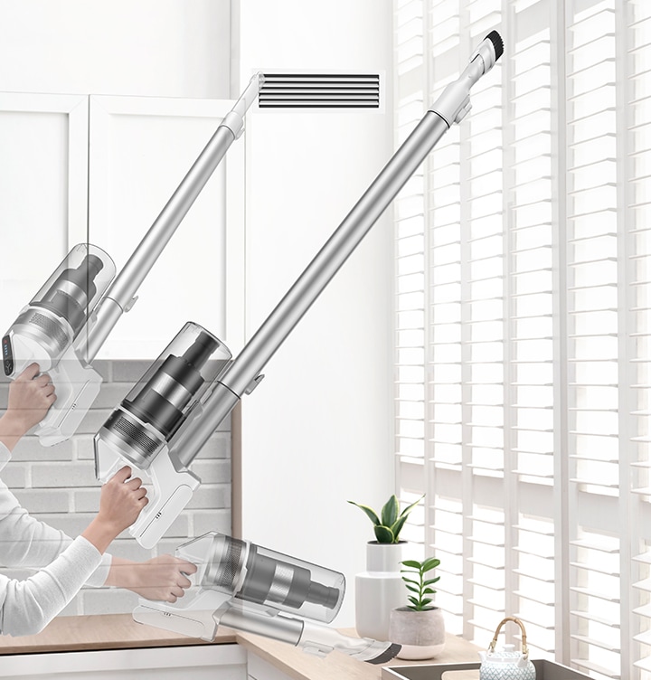 A person lifts the stick vacuum with one hand and cleans vent with the crevice tool and cleans blinds and table with the combination tool.