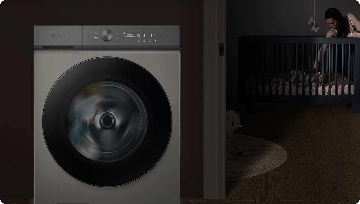 A woman in a darkened room leans into a crib to touch her baby. To the left is a wall, in front of which is a white Samsung Bespoke Grande AI washer with laundry spinning inside.