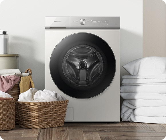 A white Bespoke Grande AI washer is in a laundry room. Three baskets of laundry are placed to the left of the machine, and folded white bedding is placed to the right of the machine.