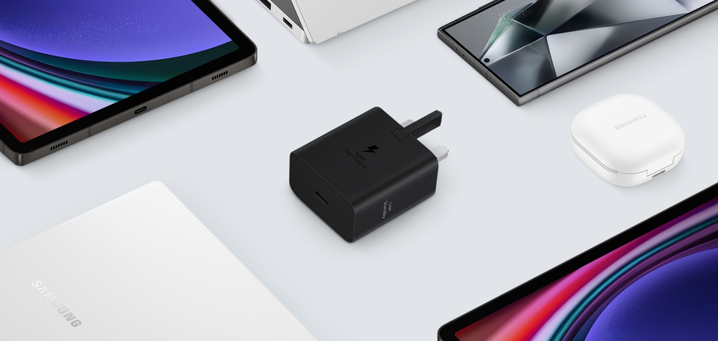 A top view of various Samsung Galaxy devices are shown with a black Power Adapter placed at the center. To the left, a Galaxy Book3, to the right Galaxy Buds2 Pro in White, a Galaxy Tab S9 and Galaxy S24 Ultra are also shown.
