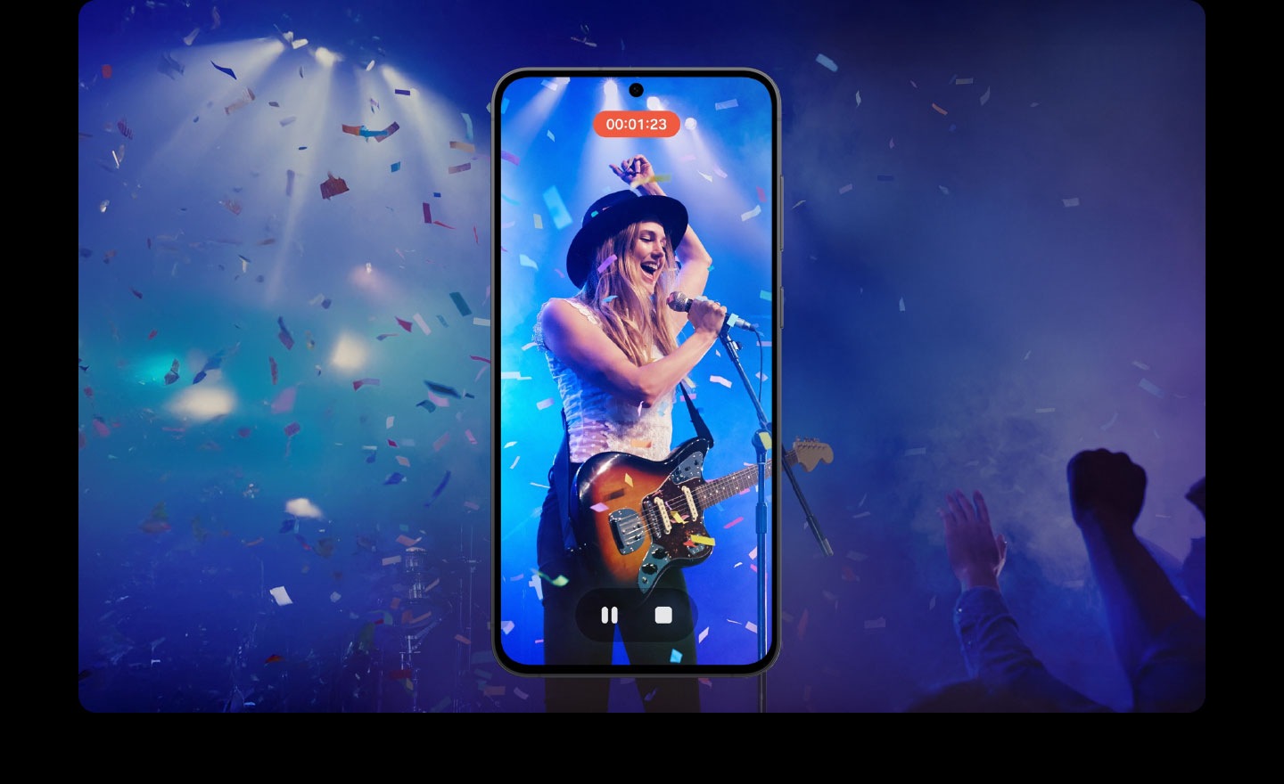 A smartphone screen shows a video recording of a live stage performance. The performer wears a black hat, plays an electric guitar, and sings into a microphone. Blue stage lights illuminate the background, with confetti falling. A timer reads '00:01:23' and playback controls are visible.