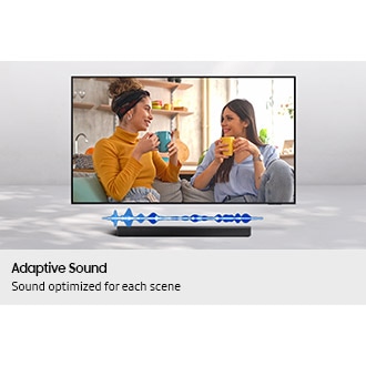 Two women, one in a cheerful yellow sweater and the other in a vibrant blue top, smile and chat while holding mugs on a cozy couch. The image features "Adaptive Sound," showcasing audio optimisation from the Samsung B650f soundbar.