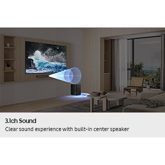 A modern living room showcases a large TV displaying a stunning crashing wave scene. Below, a sleek Samsung B650f soundbar with a built-in center speaker enhances the audio experience. A cozy seating area features a coffee table,