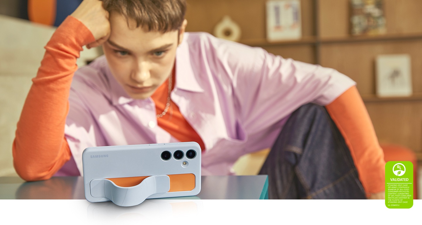 Une jeune étudiante soutient sa tête de la main tout en regardant l’écran de son Galaxy S24 Plus dans un étui ergonomique vertical Bleu Clair. La sangle forme un support qui aide le téléphone à se tenir debout, illustrant une situation pratique où la sangle est fixée pour visualiser confortablement l’écran. Le texte indique que L’ÉTUI ERGONOMIQUE VERTICAL CERTIFIÉ UL (EF-GS921) CONTIENT AU MOINS 20 % DE MATIÈRES RECYCLÉES POST-CONSOMMATION, AVEC 39 % DE MATIÈRES RECYCLÉES POST-CONSOMMATION DANS LE PLASTIQUE DE L’ÉTUI ERGONOMIQUE VERTICAL. UL.COM/ECV