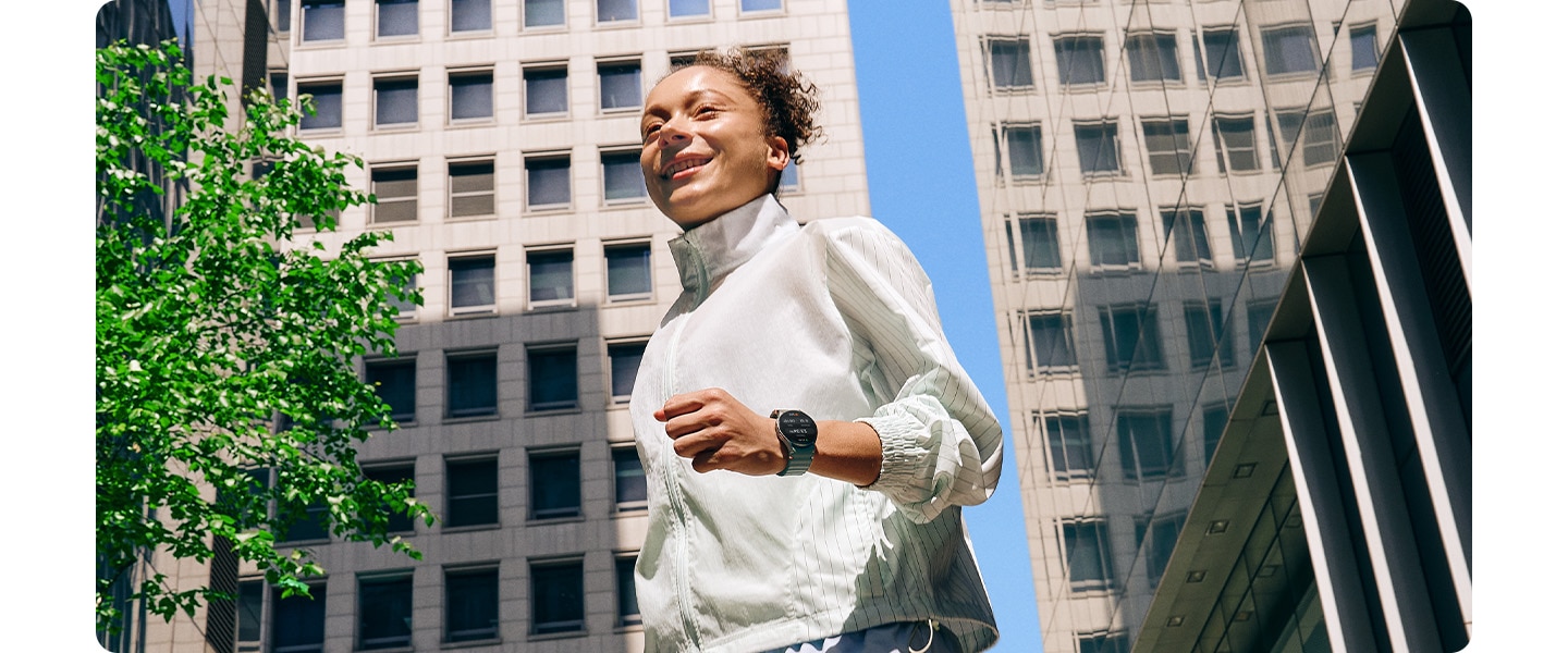 Une femme en tenue de sport portant une Galaxy Watch7 marche dans un quartier urbain entouré de bâtiments.