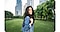 Portrait mode is on. A woman is standing in an urban park, smiling at the camera. The background, which includes grass, trees, and high rise buildings, is blurred slightly so she stands out.