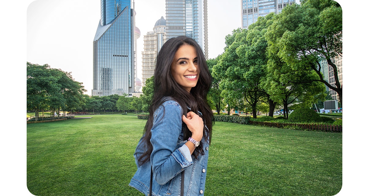 Portrait mode is off. A woman is standing in an urban park, smiling at the camera. Behind her, there is grass and some trees, and high rise buildings.