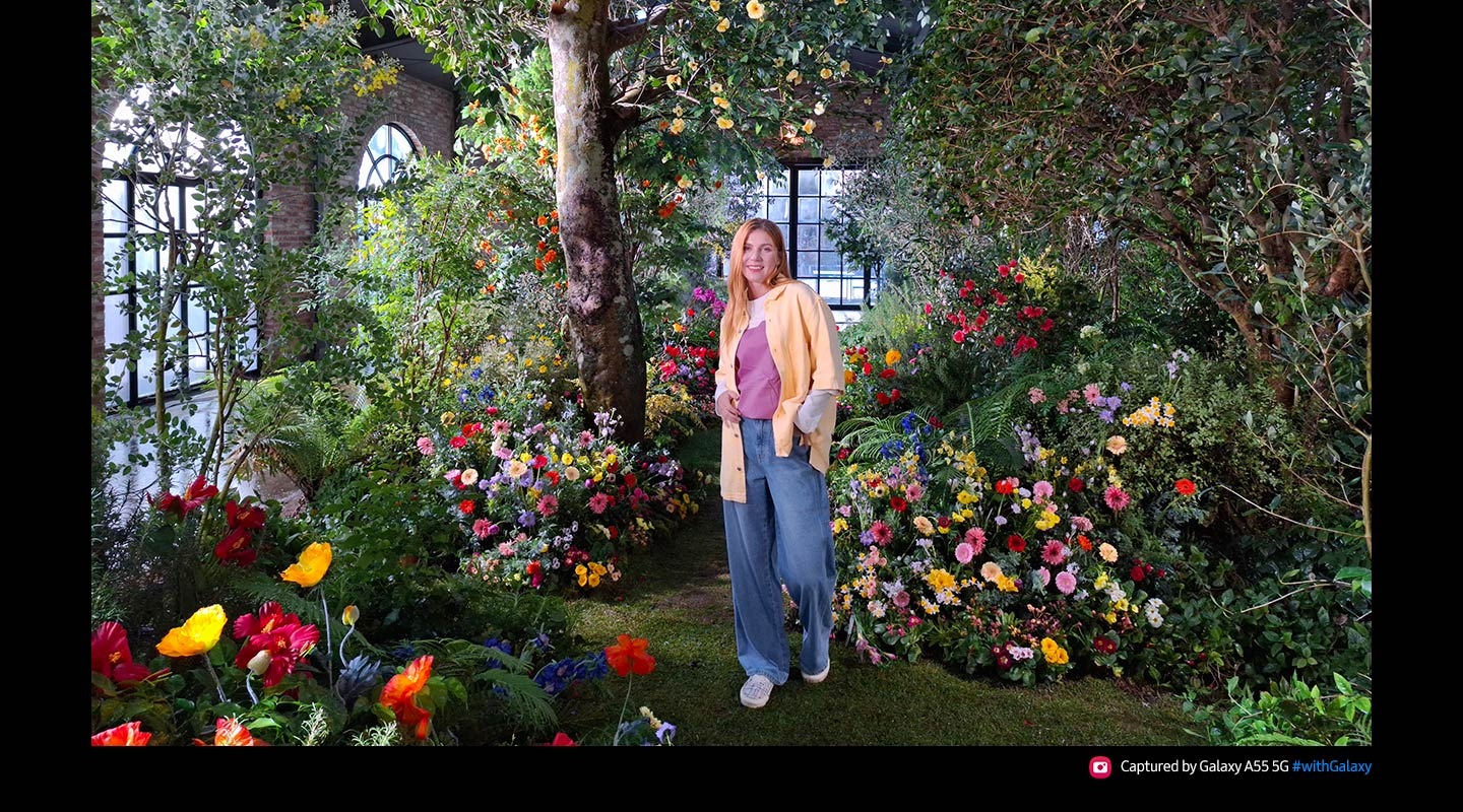 A photo taken with 50 Megapixels of high-resolution of a person standing in a lush indoor garden full of colorful flowers and greenery. Text reads Captured by Galaxy A55 5G #withGalaxy.