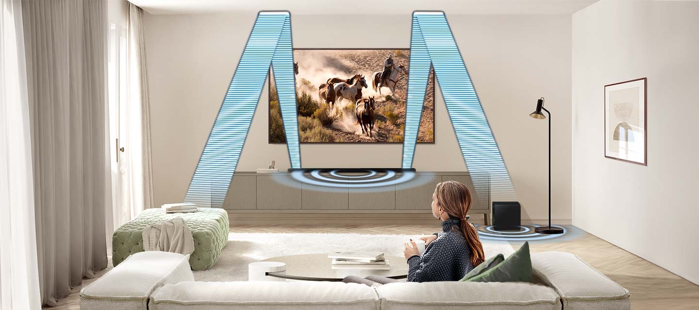 In a living room, a woman sits on a sofa and watches TV. The TV is installed onto the wall, with an Ultra Slim Soundbar and subwoofer underneath. The Soundbar gives off angled sound waves that travel up, hit the ceiling, and bounce down to the back of the room where the woman is sitting. Then round sound waves emanate from both the Soundbar and subwoofer.