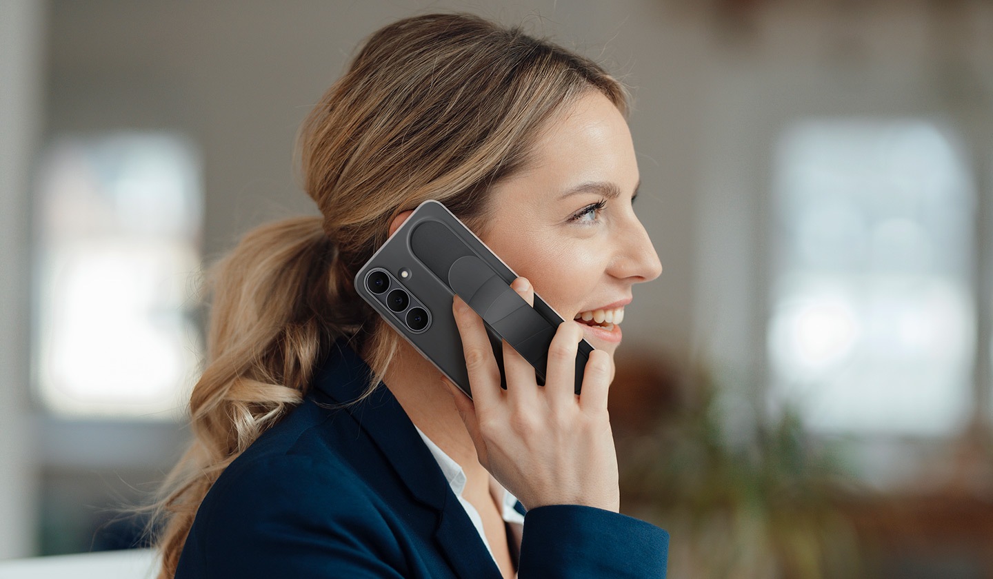 A woman is talking on her Galaxy S25 FE and has it held up to her right ear. The Galaxy S25 FE is encased in the Standing Grip Case in Black. Her fingers holding the phone is tucked underneath the adjustable grip.