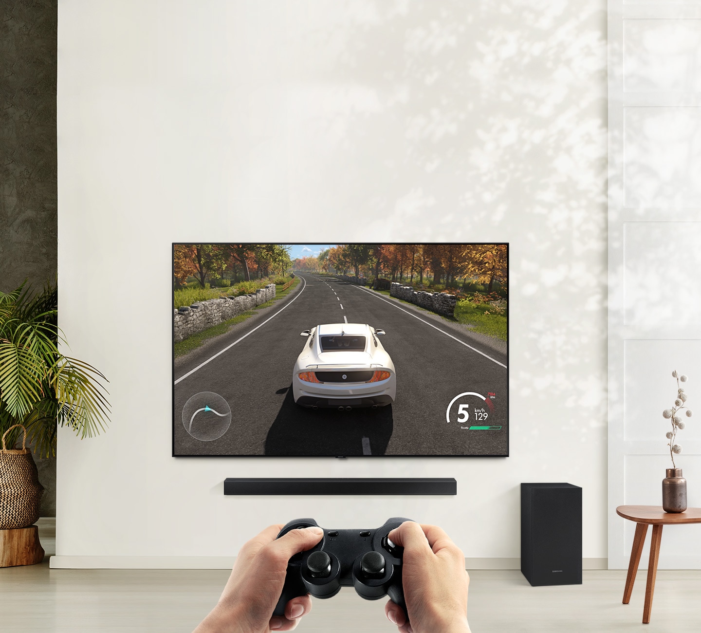 A TV hangs on a wall with a car racing game on screen. An A450 Soundbar hangs below with it's matching subwoofer on the floor nearby.