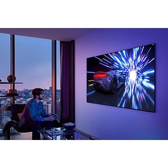 A man with a controller smiles as he games on a TV on the wall. The TV displays a racecar speeding through a lighted tunnel.