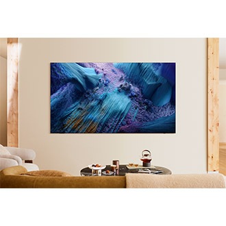A 98-inch Samsung Neo QLED TV is installed on the wall of a living area. It displays a blue abstract landscape on screen.