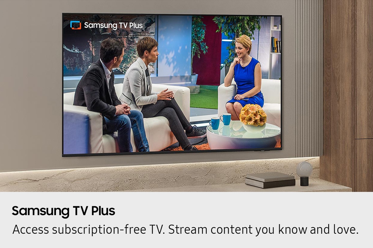 Samsung Mini LED TV mounted on a modern living room wall displaying Samsung TV Plus content, with three people seated in a studio interview setting on screen.