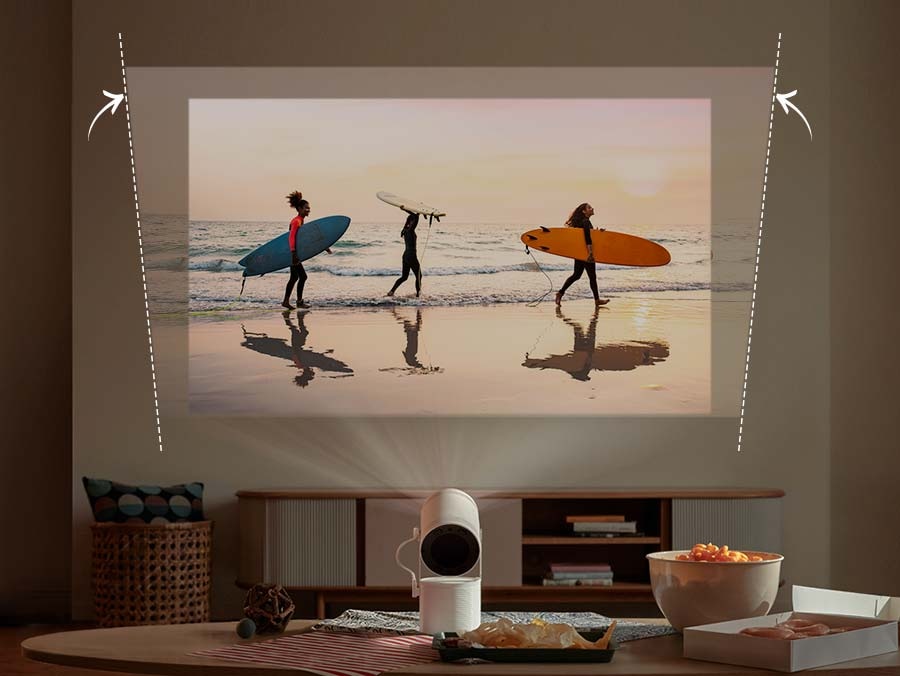 The Freestyle is projecting an image of three people carrying surfboards at a beach onto a slanted wall. The image is slanted due to the wall, but The Freestyle automatically adjusts it to make it level.