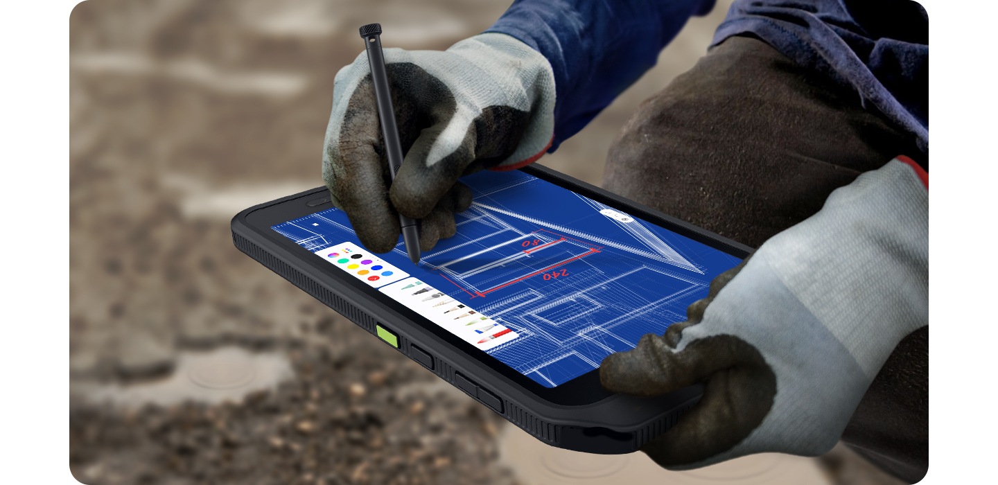 Se muestran las manos de un trabajador que usa guantes de alta resistencia, dibujando un diseño arquitectónico en un dispositivo Galaxy Tab Active5 5G con un S Pen. El fondo muestra terrenos difíciles.