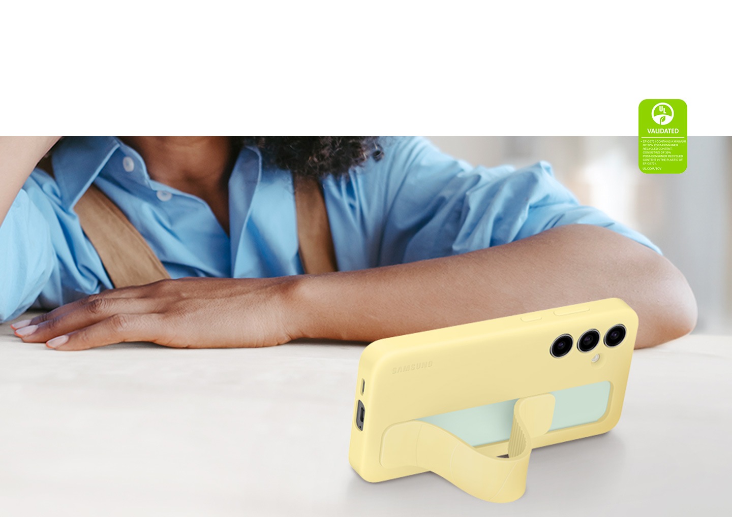 Una mujer apoya el brazo sobre una mesa mientras mira algo en un Galaxy S24 FE con una cubierta de agarre vertical en Yellow. La correa actúa como un soporte, colocando el dispositivo horizontalmente en un ángulo de visión cómodo. El texto dice: CUBIERTA DE AGARRE VERTICAL VALIDADA POR UL (EF-GS721) CONTIENE UN MÍNIMO DEL 22 % DE MATERIAL RECICLADO POSCONSUMO, QUE SE COMPONE DE 39 % DE MATERIAL RECICLADO POSCONSUMO EN EL PLÁSTICO DE EF-GS721. UL.COM/ECV