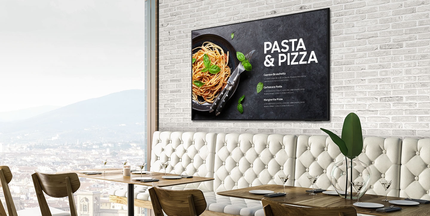 Un producto con pantalla de diseño delgado está colgado en la pared de un restaurante con un fondo interior blanco que muestra el paisaje urbano a la izquierda. El producto muestra fotos de la comida, que incluye pasta carbonara y pizza margarita.