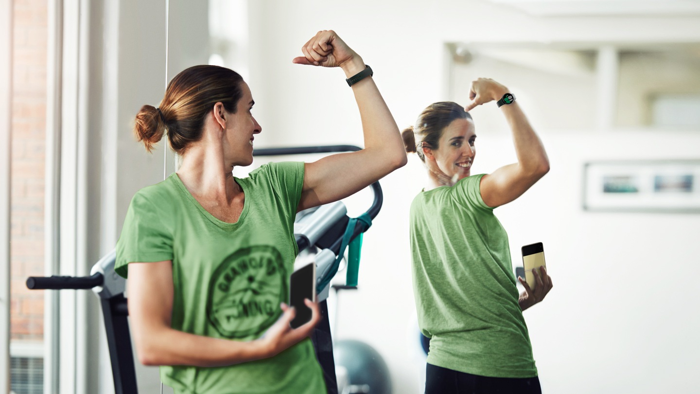 A woman is looking at the mirror and flexing her muscles while wearing a Galaxy Watch4 device and holding a Galaxy Z Flip3 5G in her right hand.