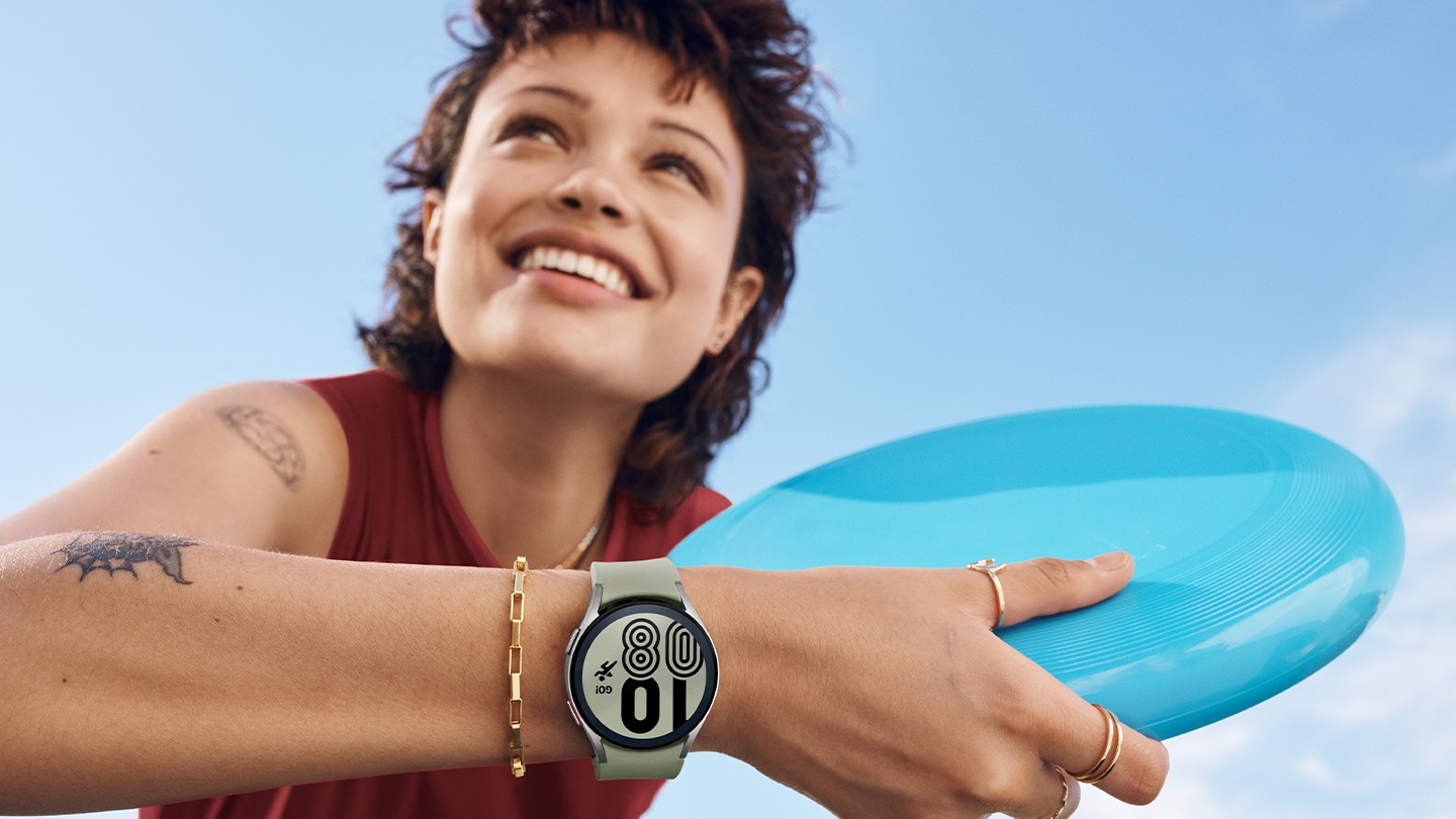 A woman is smiling and holding a frisbee while wearing a silver Galaxy Watch4 on her wrist.