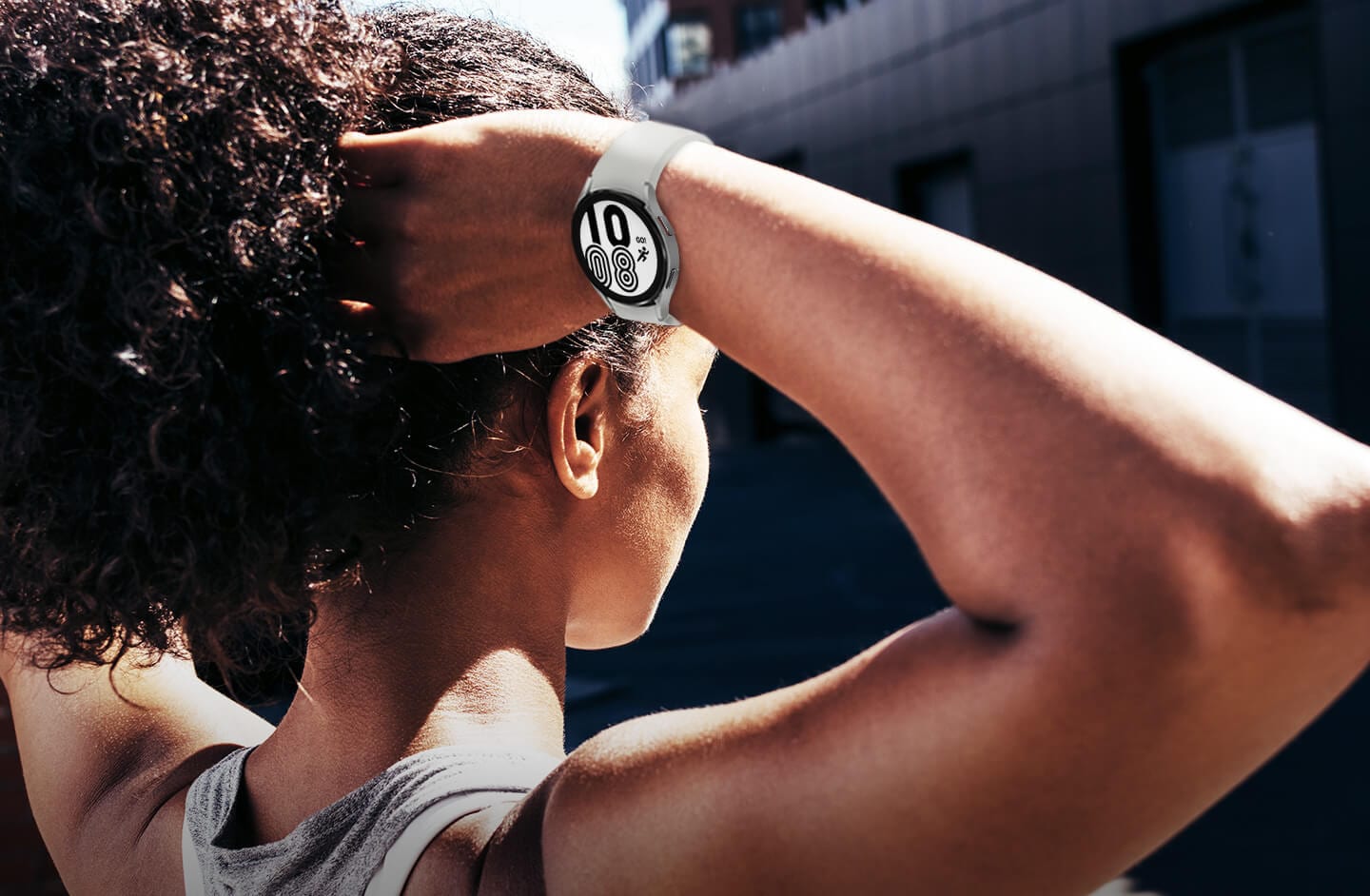 There is a woman outside wearing workout clothes and raising her hands to tie her hair. She is wearing a Galaxy Watch4 with a Silver color sport band. The watch is displaying the time and a UI icon with a running symbol that says "GO!" above.