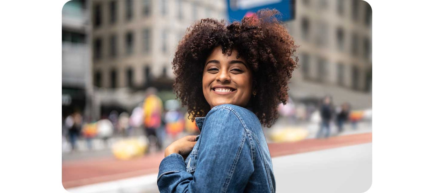 A smiling woman is shown in the center of an urban setting with the background, showing buildings and people, blurred behind her.