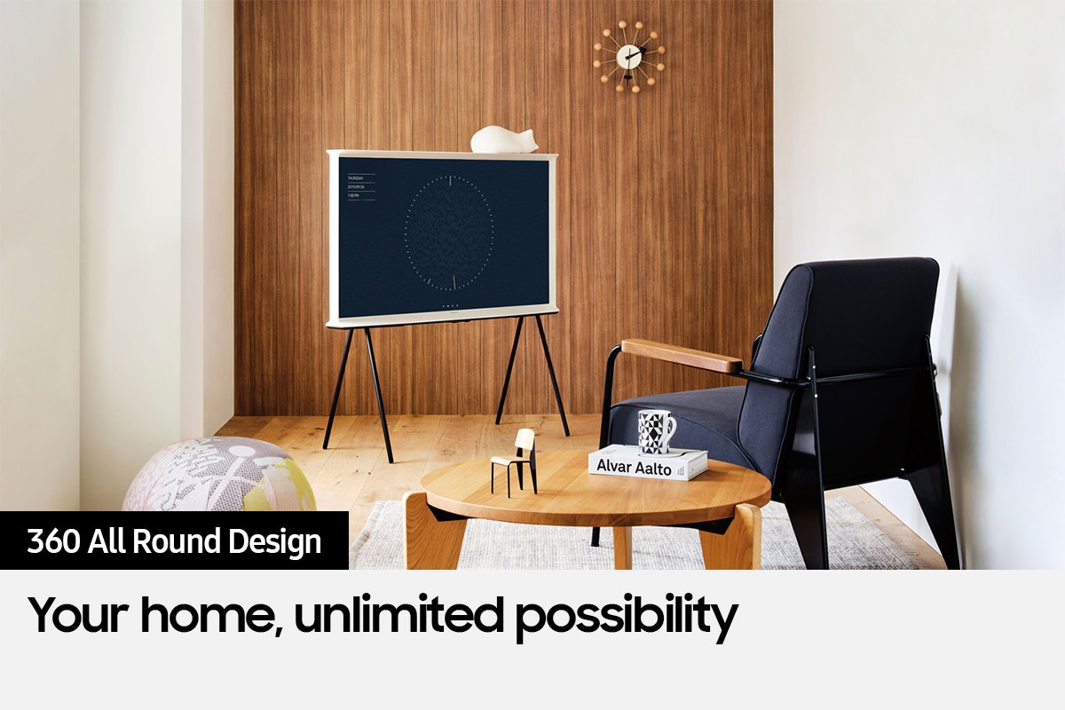 The Serif is on a stand next to a chair and small table.