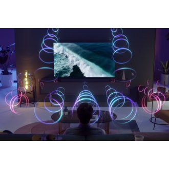 A person is sitting in front of a QLED TV as sound waves fill the room with 3D surround sound.