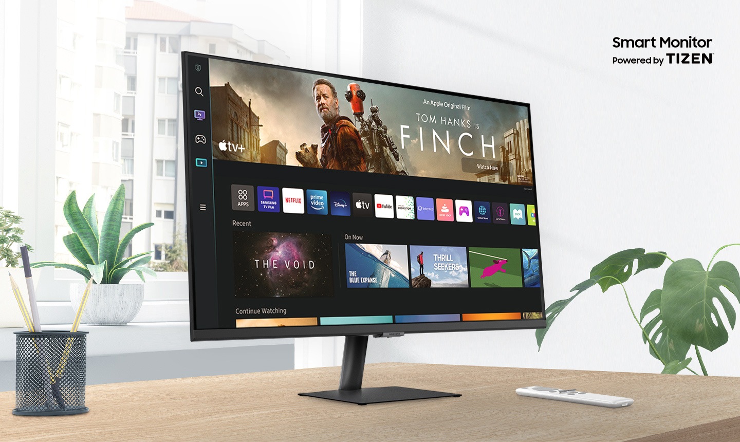 'The monitor sits on top of a wooden desk in front of a window surrounded by plants. The words 'Smart Monitor Powered by Tizen' are shown above the monitor. The screen shows a variety of content apps and individual content choices in a card format. A remote rests next to the monitor.
