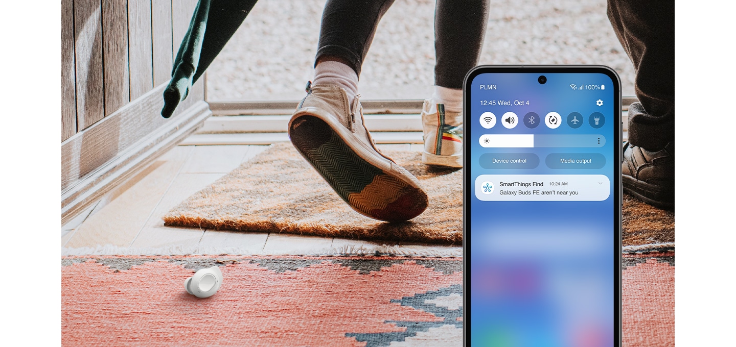 A smartphone display with the app SmartThings on. A person is seen walking out of a location and one Galaxy Buds FE earbud is left on the carpeted floor. The device's screen shows a notification message that reads Galaxy Buds FE aren't near you.