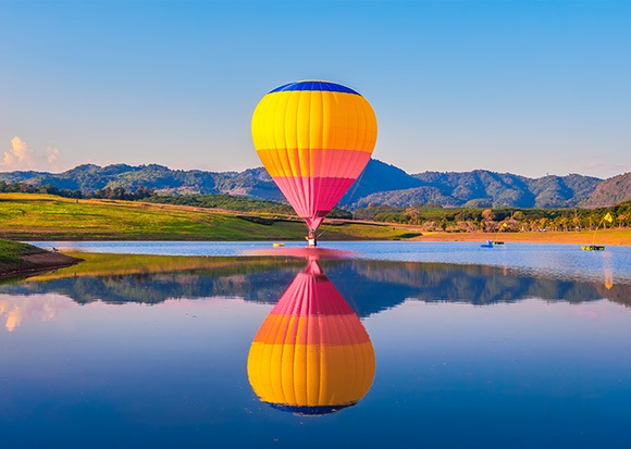 A colorful hot air balloon floating above a lake, with its reflection on the water.