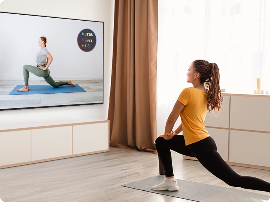A woman exercises while watching a fitness trainer on TV. Her workout stats from Galaxy Watch appear as an on-screen overlay.