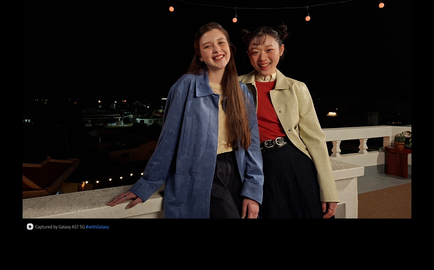 Video of two young women smiling and waving while leaning on an outdoor terrace against a night backdrop, taken with Galaxy A57 5G's camera system. The details in the video are clear and bright thanks to optimized Nightography. At the bottom is camera icon with text Captured by Galaxy A57 5G hashtag with Galaxy.