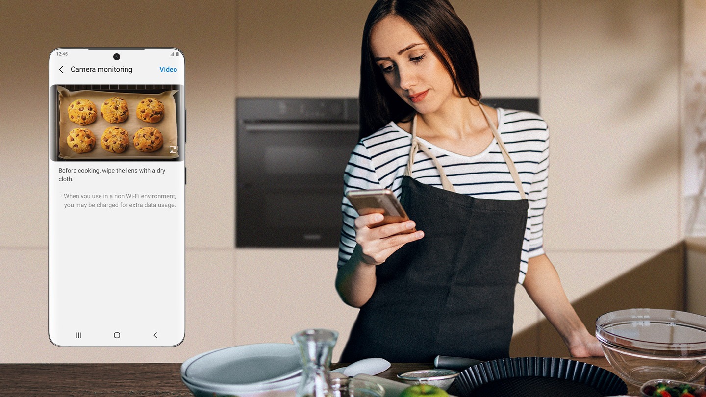 Egy nőt ábrázol a sütő előtt, és az okostelefonját nézi.  A telefon képernyőjének közeli képén a SmartThings App Camera Monitoring funkciója látható, amely valós idejű videót jelenít meg a sütőben sült süteményekről.