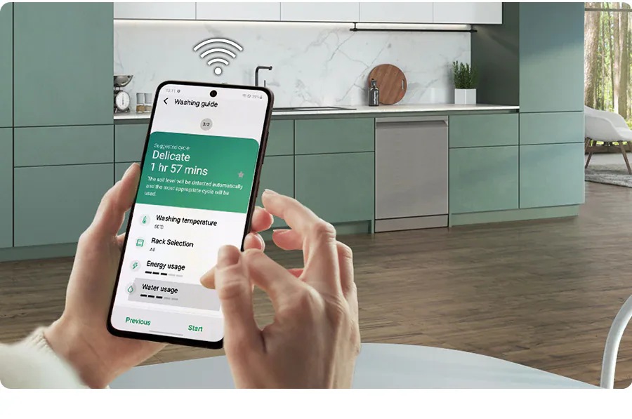 Egy okostelefon, amelyen a SmartThings alkalmazás engedélyezve van, zöld konyhai háttér előtt, látható Bespoke DW60BB890UAPET beépített mosogatógéppel