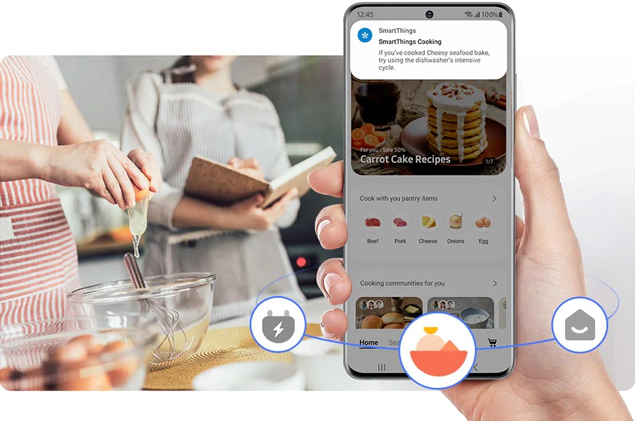 Egy kézben tartott okostelefon, amelyen a SmartThings Cooking alkalmazás látható, a háttérben két ember süteményt készít.