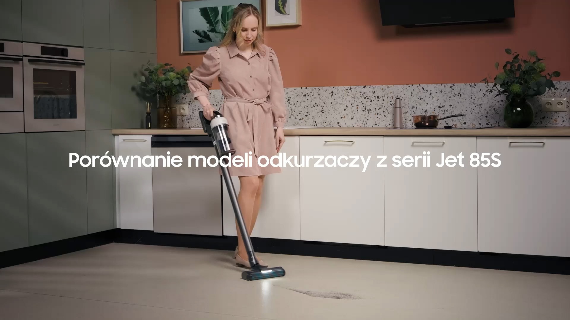 Kobieta ubrana w różową sukienkę trzyma w ręku odkurzacz Samsung serii Jet 85S i odkurza podłogę w kuchni.