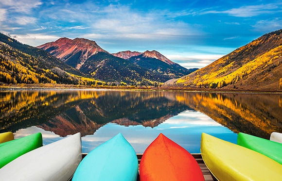 A beautiful landscape photo of the blue sky and mountains in the background, a still, reflective lake and colorful canoes in the foreground.