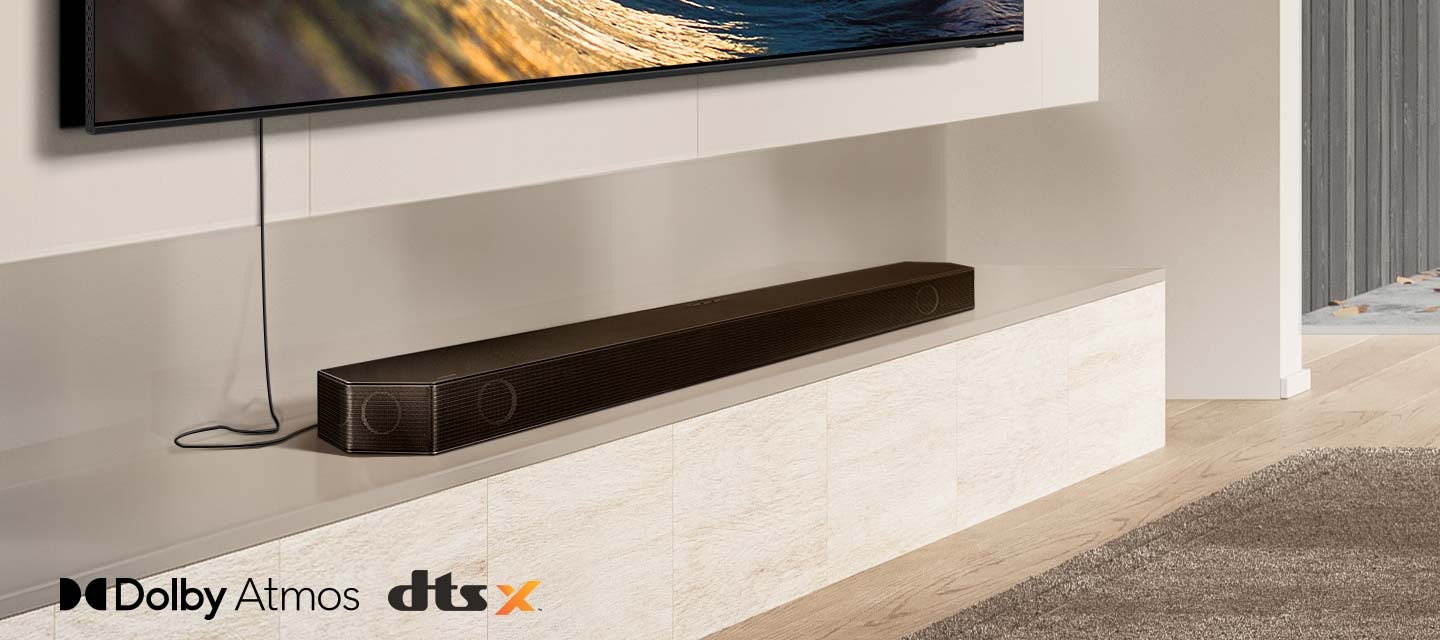 Un prim plan pe o bară de sunet așezată sub un televizor, împreună cu siglele pentru Dolby Atmos și DTS:X. O pictogramă de semnal Wi-Fi apare lângă Soundbar. Scena se micșorează pentru a dezvălui o întreagă cameră de zi, cu difuzoarele din spate instalate în spate. Undele sonore emană din bara de sunet și din difuzoarele din spate, reflectându-se pe tavan și călătorind în jurul camerei.
