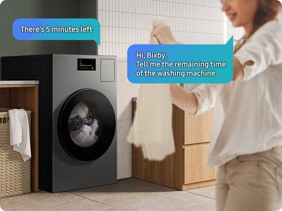 A woman in a laundry room asks Bixby the remaining time in the cycle, "Hi bixby. Tell me the remaining time of the washing machine.". Bixby replies from the Laundry Combo, "There's 5 minutes left.". 