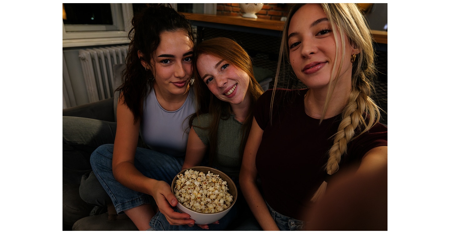 Trei fete pe o canapea, acasa, intr-un ambient slab luminat, realizand o fotografie de grup. Una dintre fete face fotografia folosind Galaxy A37 5G, cu Nightography, pentru detalii foto luminoase si vibrante.